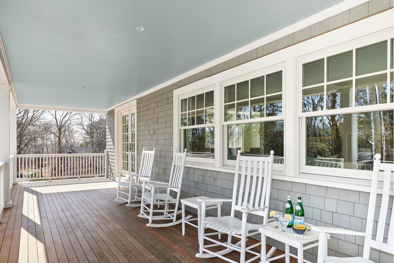 A spacious porch features white wooden rocking chairs and a small table with drinks and glasses. The ceiling of the porch is painted blue. The wooden floor, gray shingle siding, and large windows complement the view of trees.