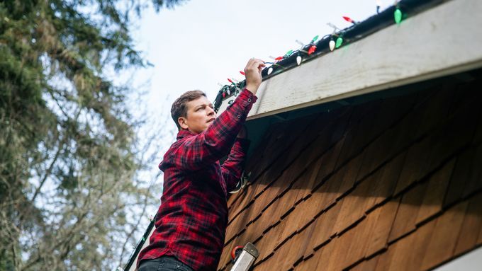 A man in a flannel shirt standing on a ladder to hang Christmas lights on his home.