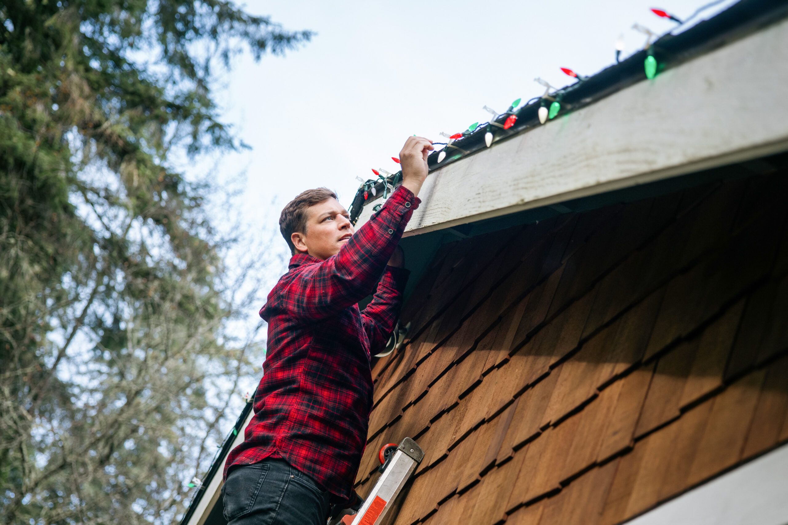 A man in a flannel shirt standing on a ladder to hang Christmas lights on his home.