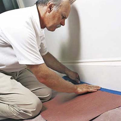 An image of a man placing tape at the base of a wall to prepare for painting.