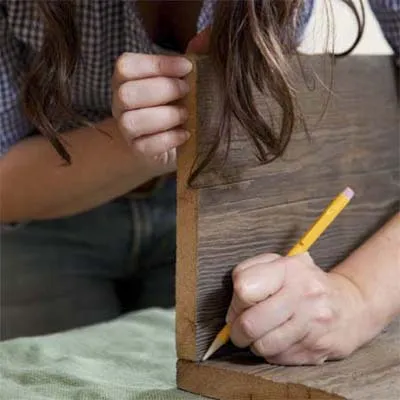 A woman marks two pieces of wood with a pencil for the towel rack shelf.