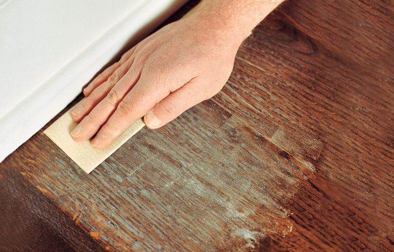 A person uses sandpaper to sand the perimeter of the room to prepare for refinishing the hardwood.