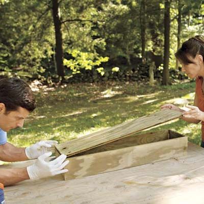 Two people attach the bottom of the frame of a window box.