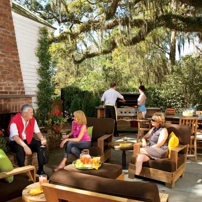 Wooden outdoor furniture with brown pillows and a grill underneath a covered spot as well as an outdoor kitchen table.