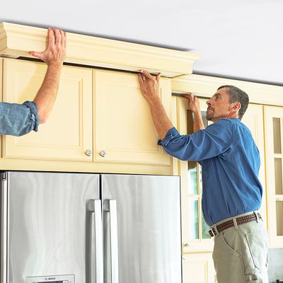 Two people install crown molding above a kitchen cabinet.