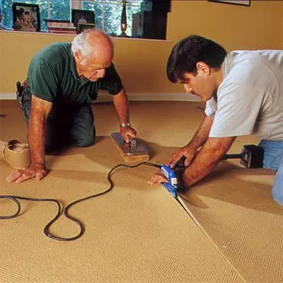 Two men seam together adjoining carpet pieces.
