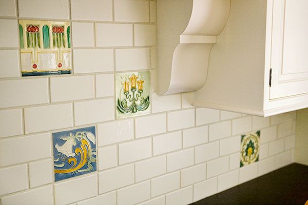 A white tile wall, with a few tiles with art on them, one has a white lily, one has a trio of candles.