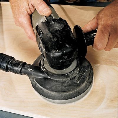 A person using a random orbit sander to sand wood.