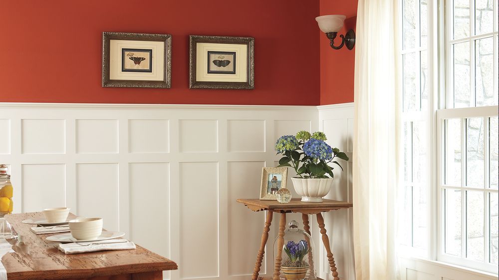 A dog sitting in the corner of a room beside the wainscotting that lines the walls.