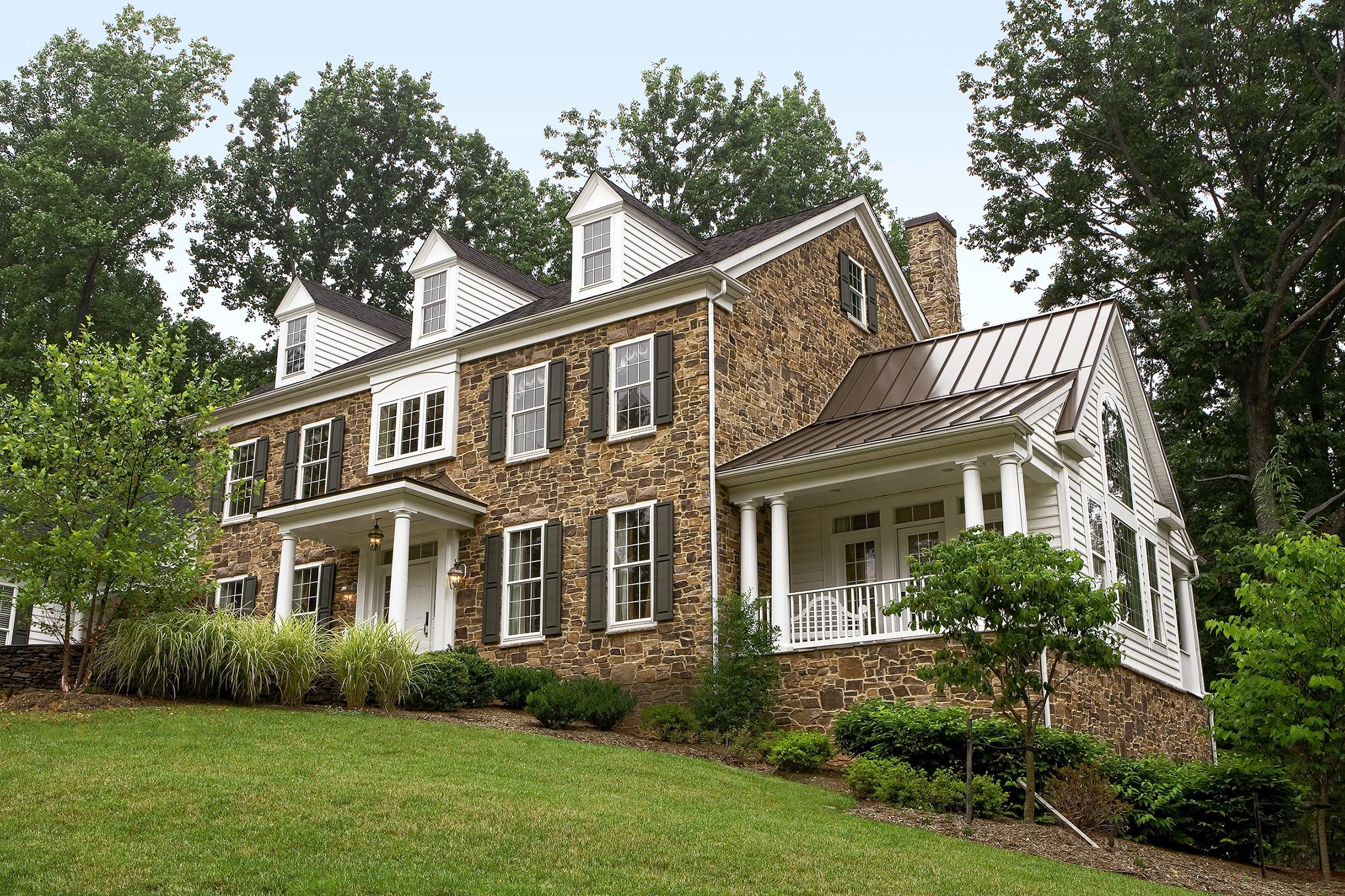 A two-story house with a stone veneer facade, white trim, a metal roof, and a large porch surrounded by greenery and trees.