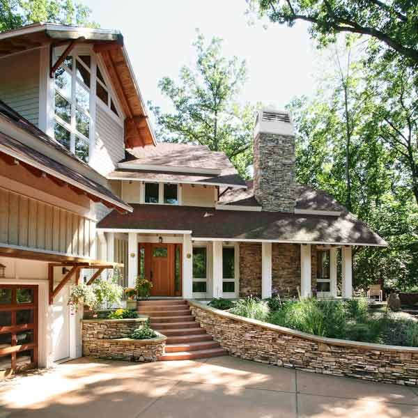 A modern house featuring a mix of stone veneer and wood siding, large windows, and a distinctive stone chimney, with a landscaped entrance and a curved retaining wall.