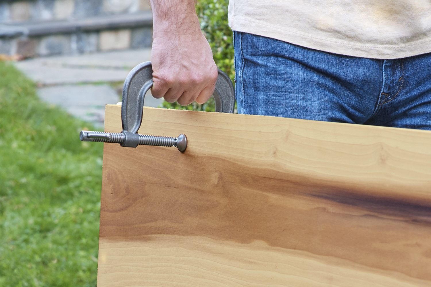 A man carrying a wooden board by affixing a C-clamp to it to use as a handle.