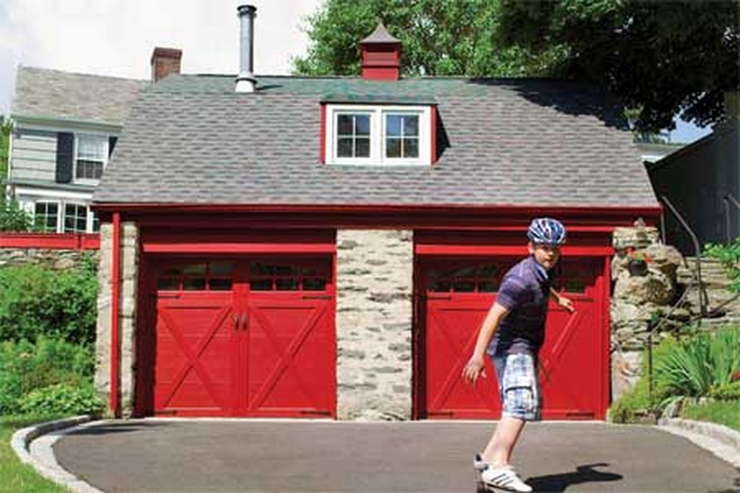 A person roller skating in front of a house with two red carriage-style garage doors.