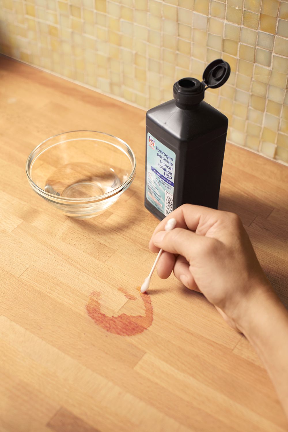 A man applying hydrogen peroxide to the surface of a butcher block counter using a Q-tip.