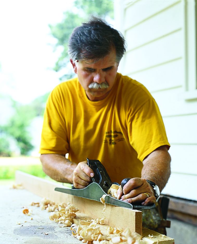 A man using a handplane on a board of wood on top of a work table.