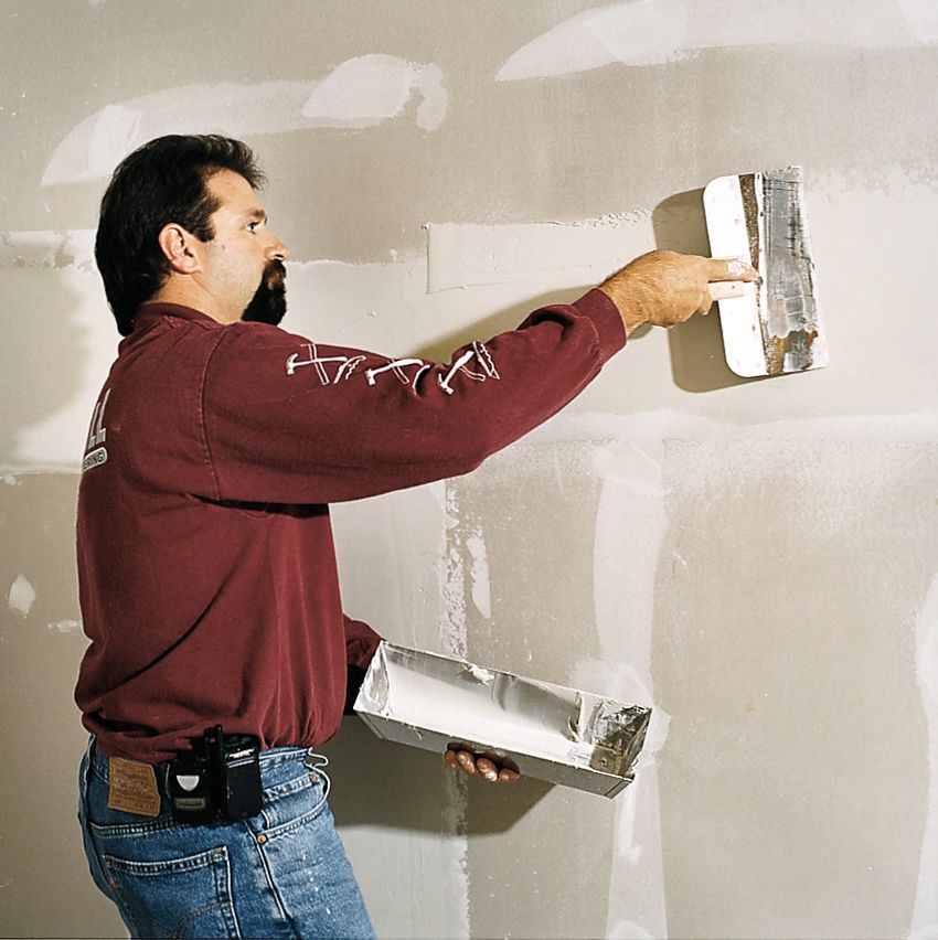 A man uses a wide drywall knife and mortar box to finish drywall.