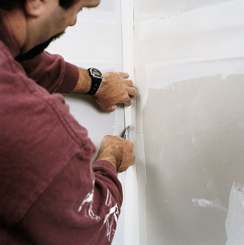 A man uses a drywall knife to push the creased tape into the corner where two drywall meet.