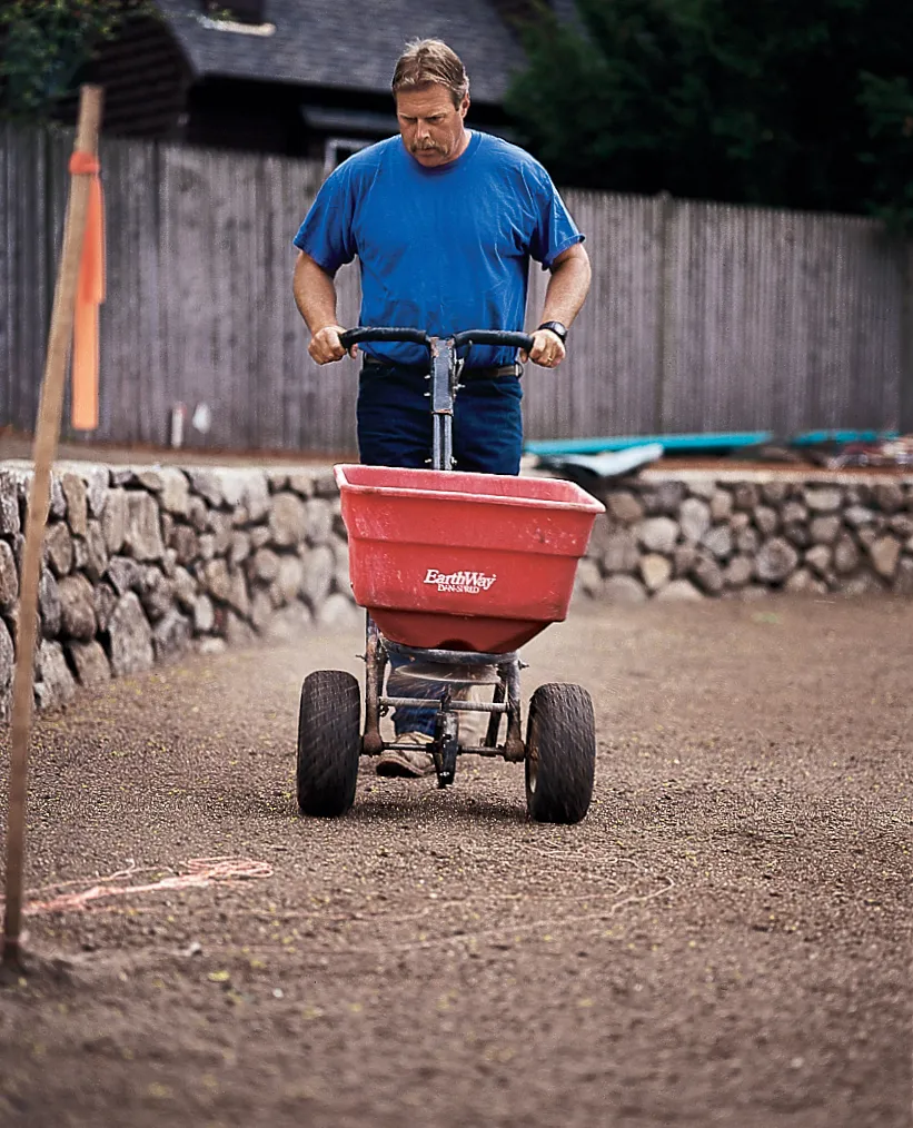 Rodger Cook uses a red fertilizer spreader on a freshly prepared soil surface.