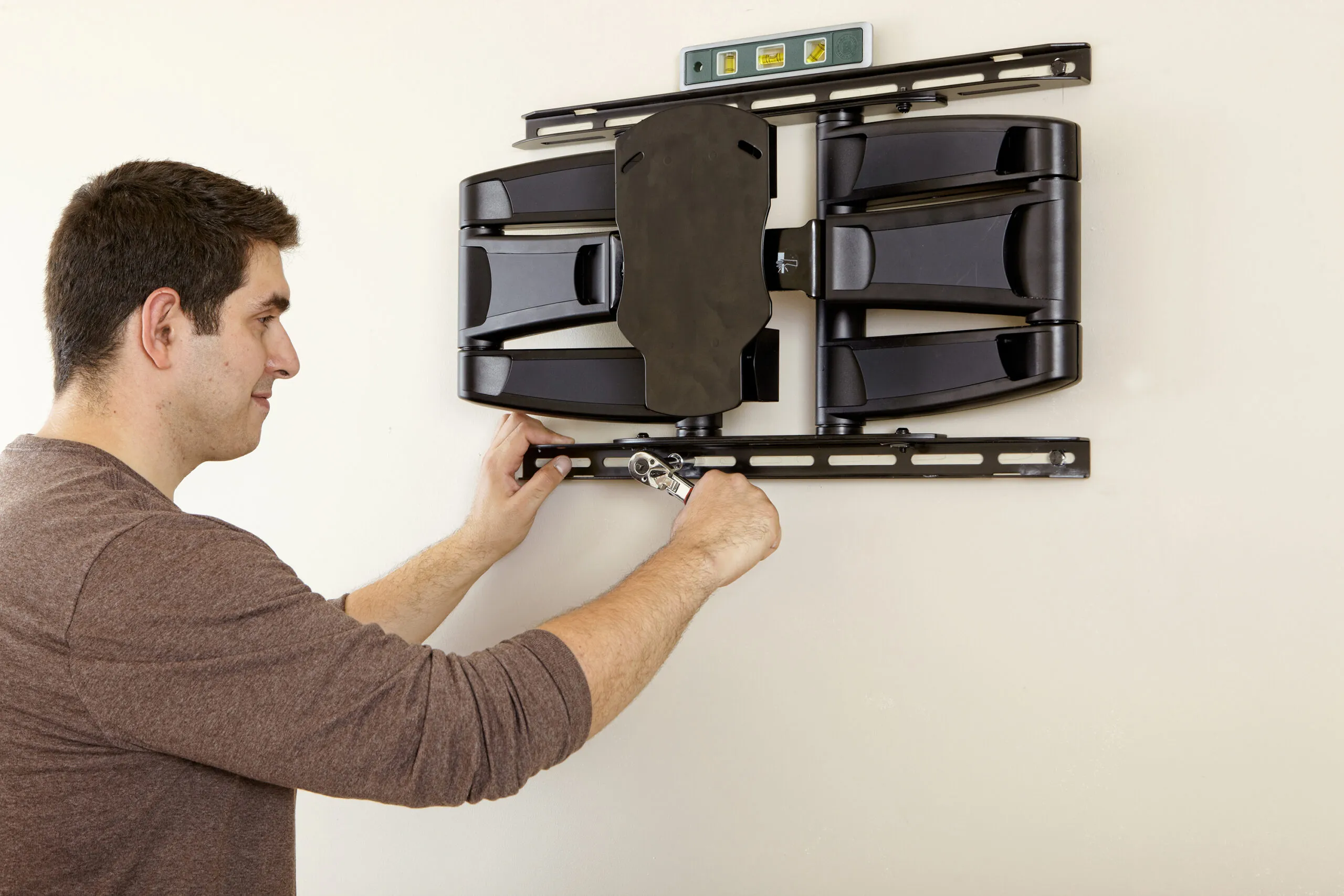 A person uses a wrench to secure a TV wall mount to the wall, with a level placed on top to ensure proper alignment.