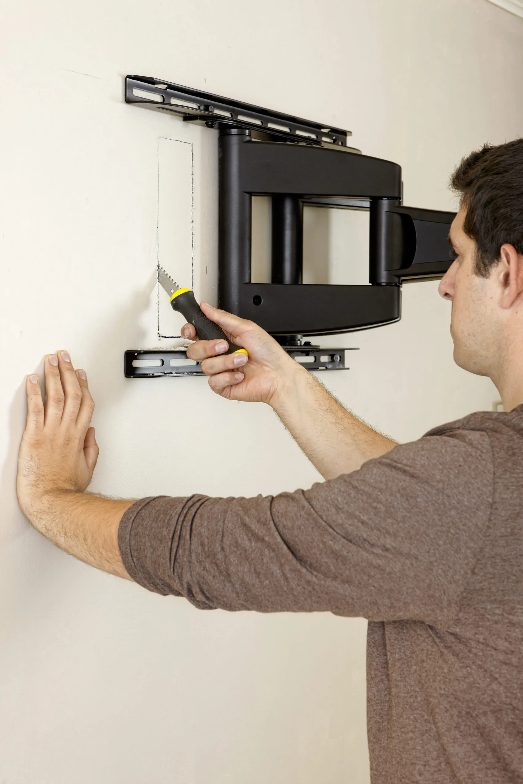 A person cutting a rectangular hole in a wall using a drywall saw, next to a TV wall mount, in order to insert a cable management system.