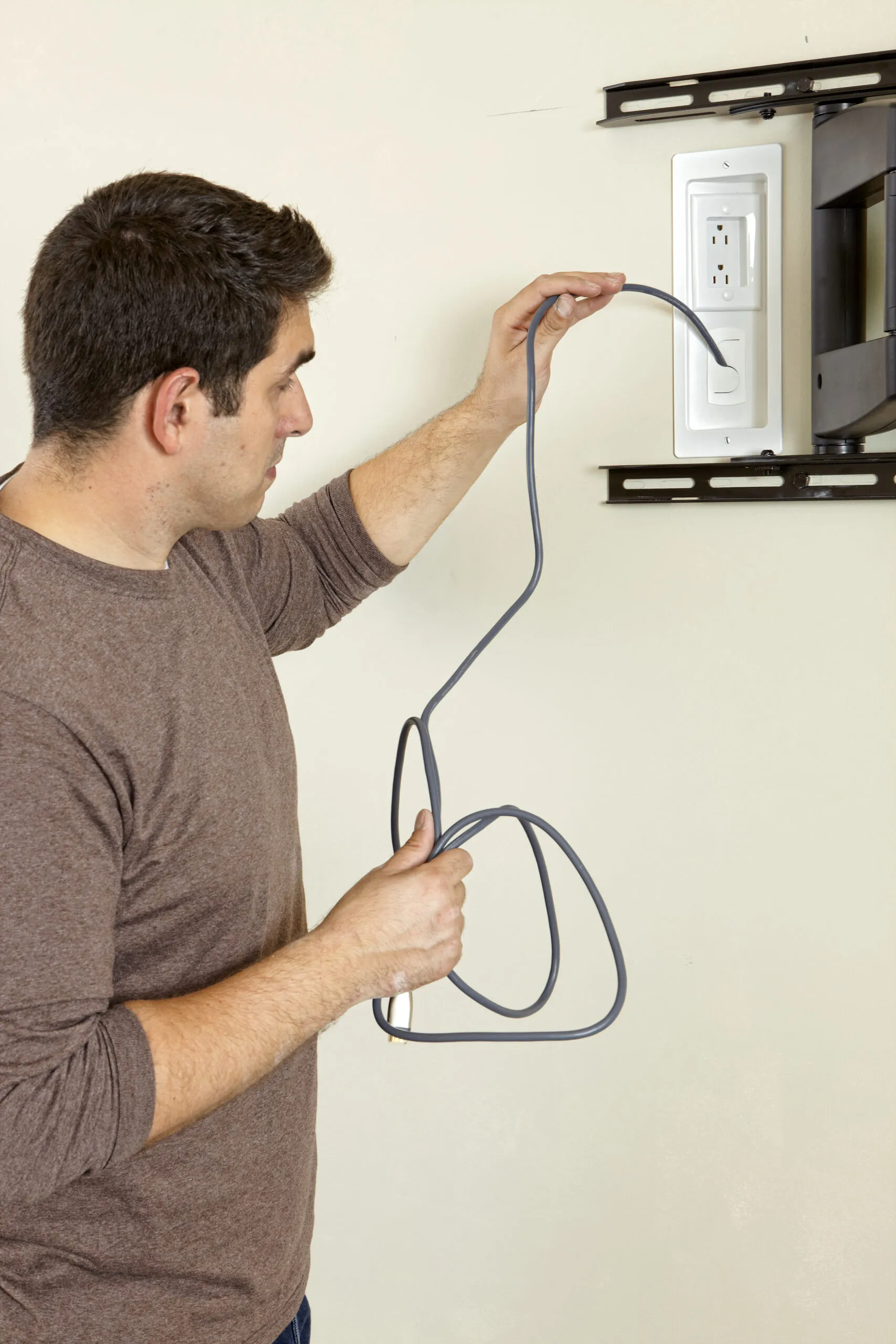 A person holding a cable and preparing to connect it to a recessed power outlet installed in a wall next to a TV mount.