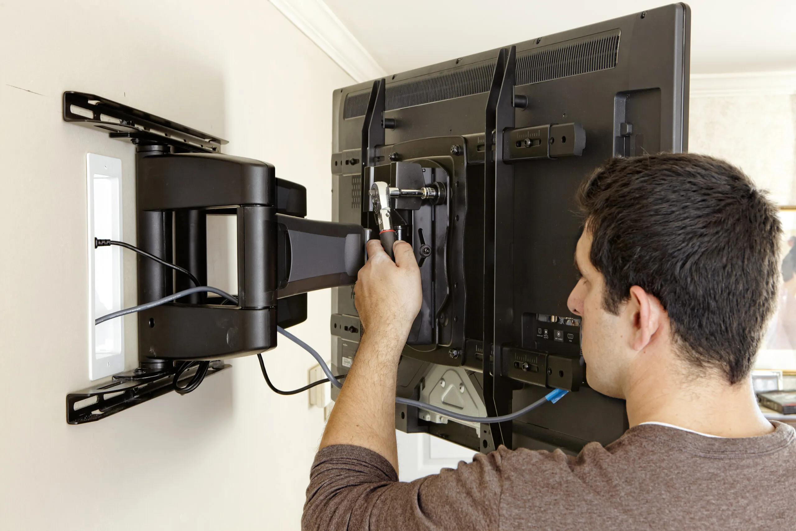A person hanging and securing a TV to a wall mount with a wrench while connecting cables to the back of the TV.
