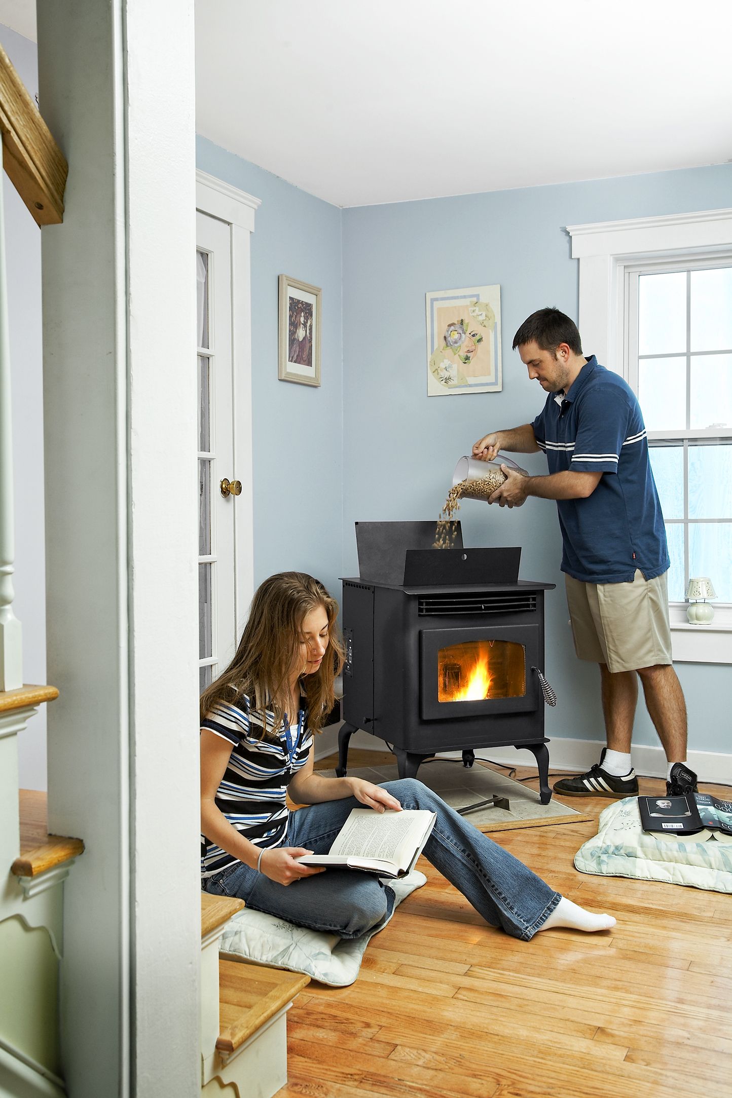 A man adds pellets to a pellet stove while a woman reads.