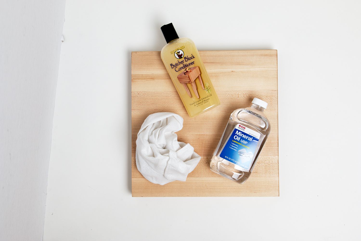 A tile of butcher block countertop with a rag, a bottle of mineral oil, and a bottle of butcher block conditioner.