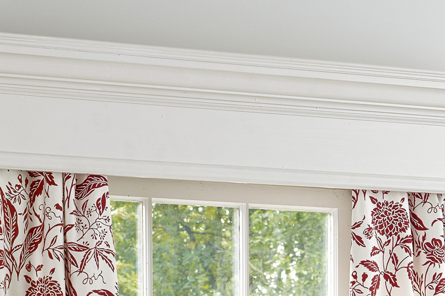 A indow cornice with whit curtains hanging from it. The curtains have a red, floral pattern.