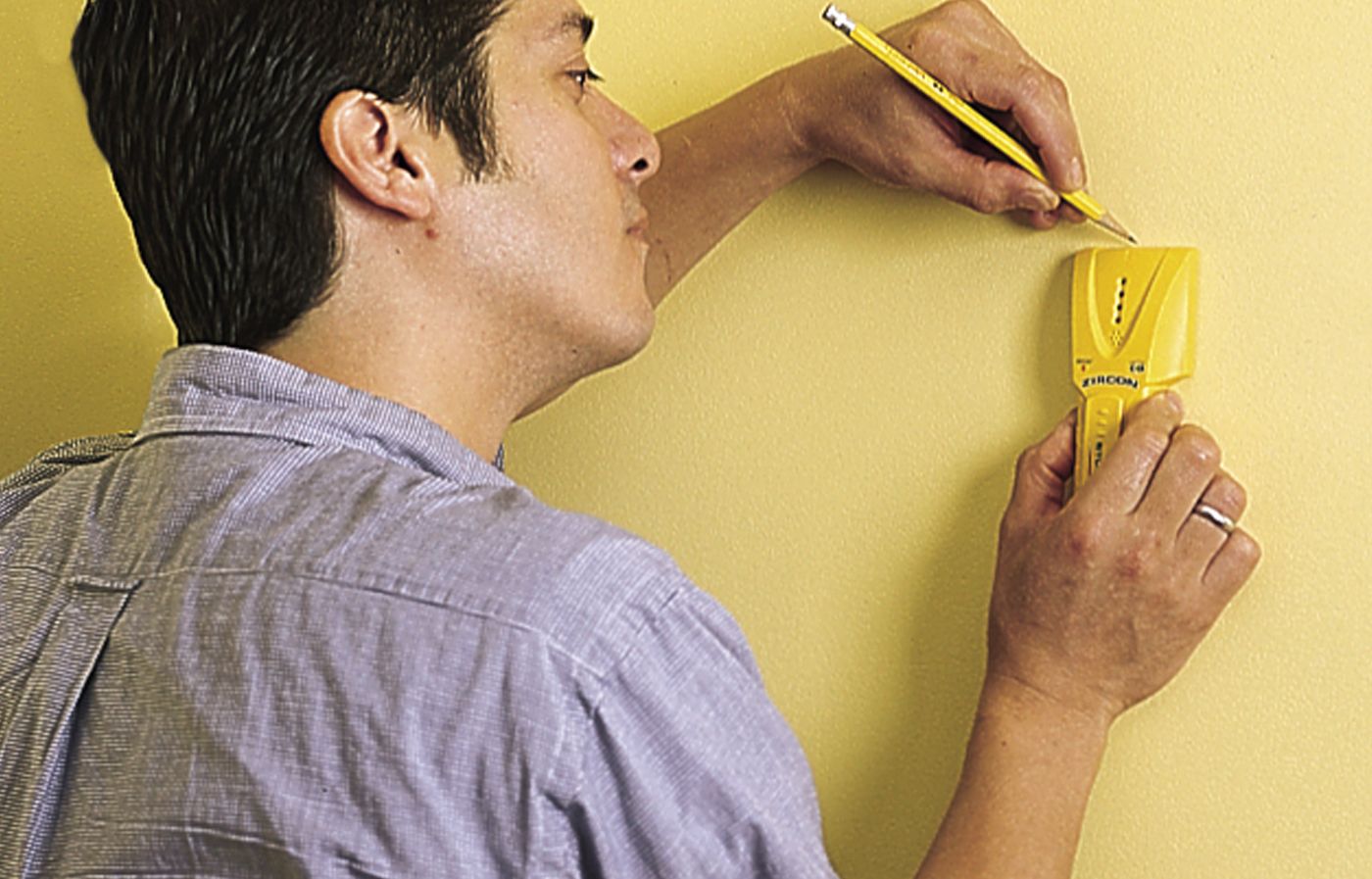 A person using a stud finder and marks the stud location with a pencil in preparation for hanging wall-mounted shelves.