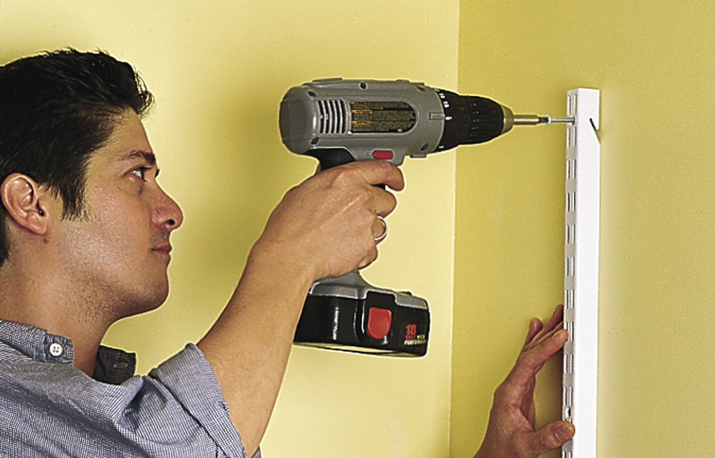 A person drills a shelf standard into the pilot hole as a step in hanging wall-mounted shelves.