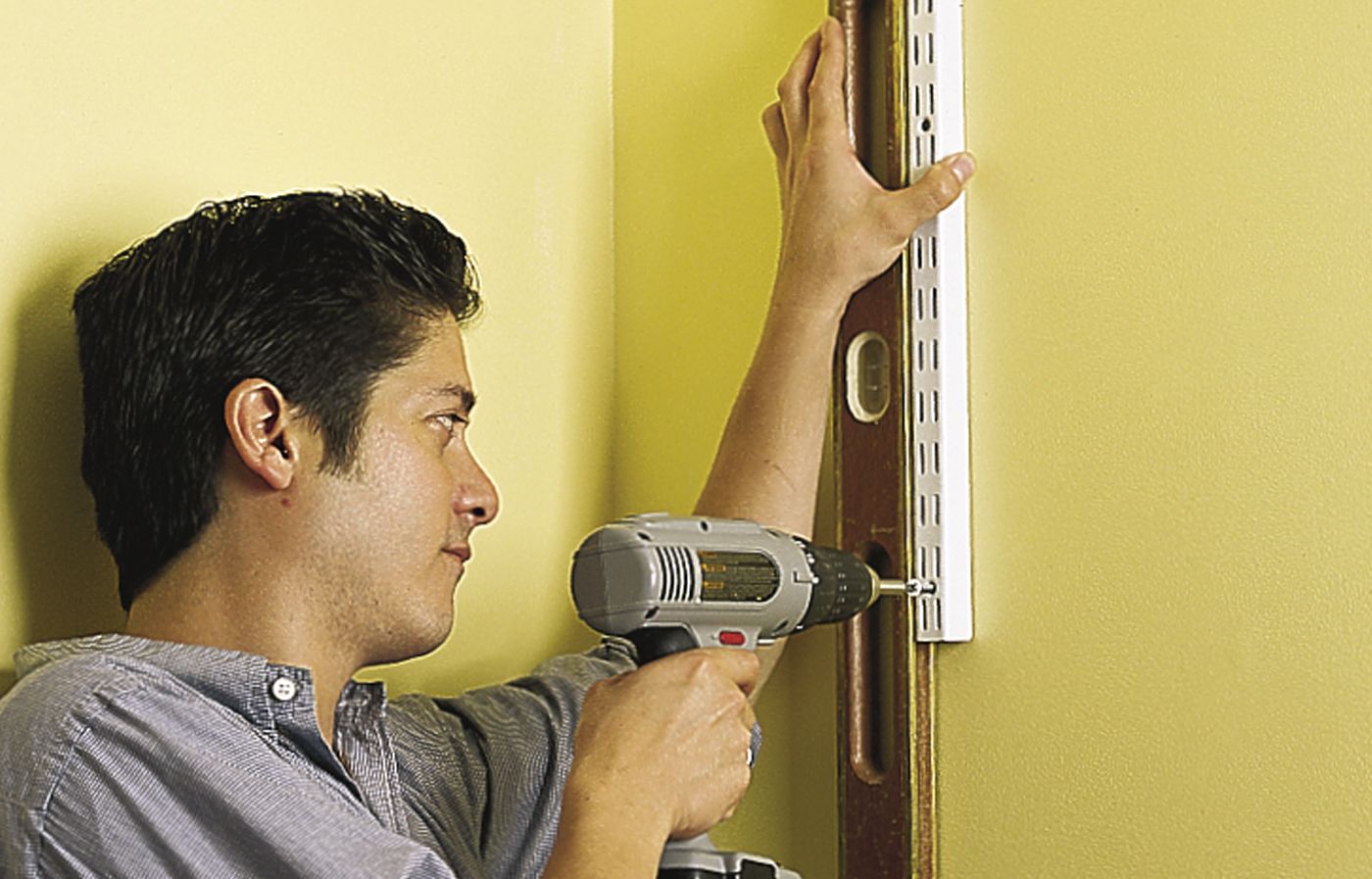 A person drills the bottom screw of a shelf standard into the wall as a step in hanging wall-mounted shelves.