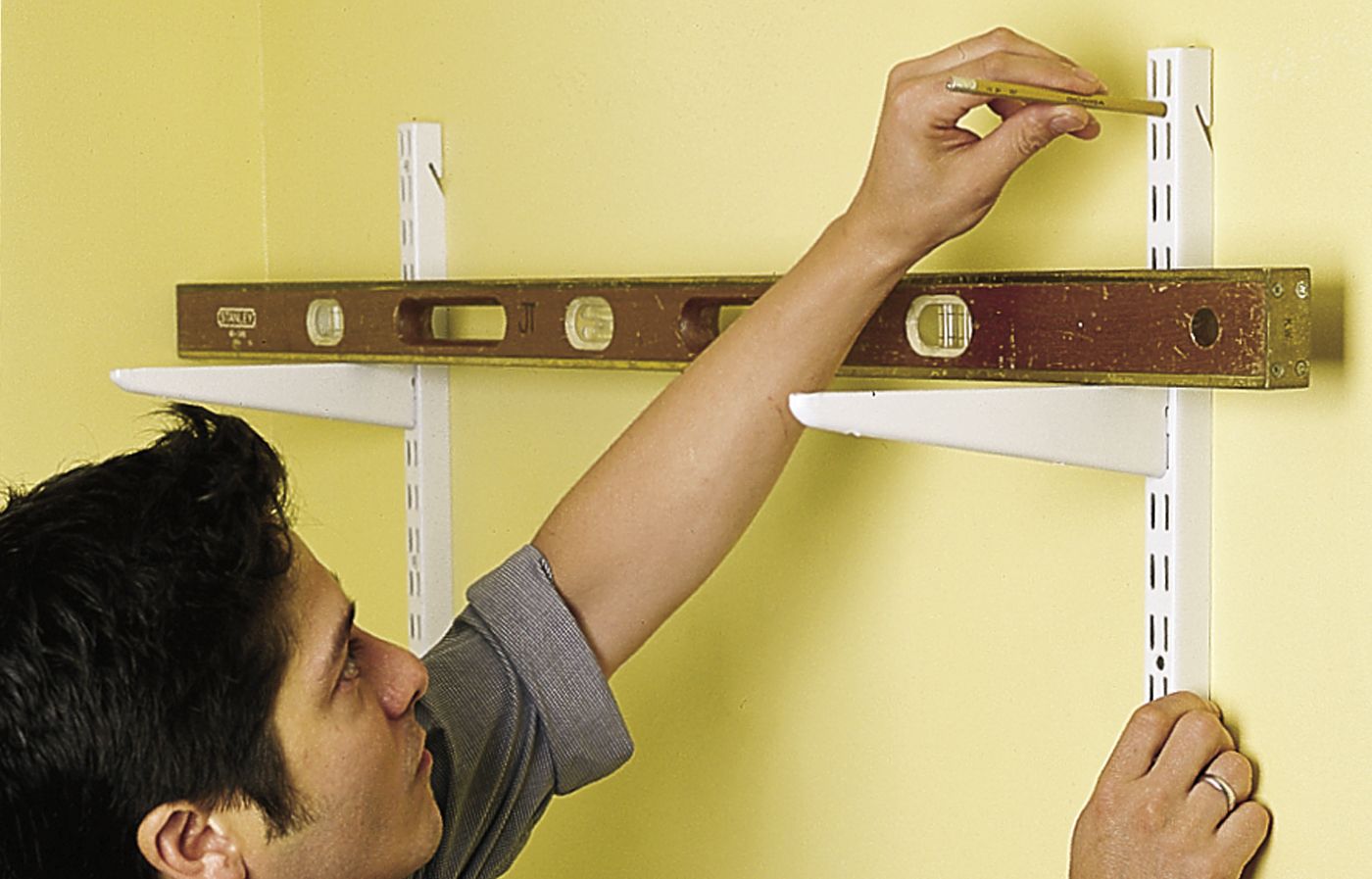 A person uses a level and marks hole with a pencil before hanging the second shelf standard as a step in hanging wall-mounted shelves.