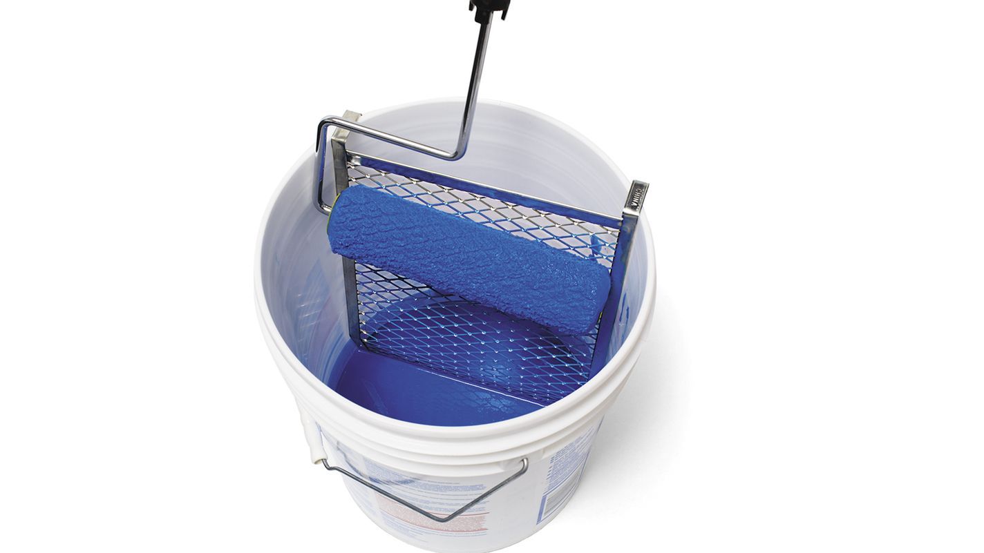 A paint roller is seen resting on a metal grid inside a white bucket, filled with blue paint.