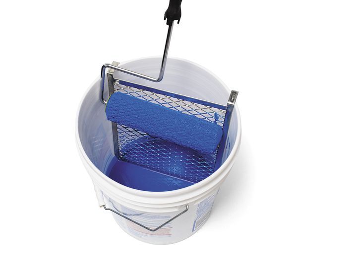 A paint roller is seen resting on a metal grid inside a white bucket, filled with blue paint.