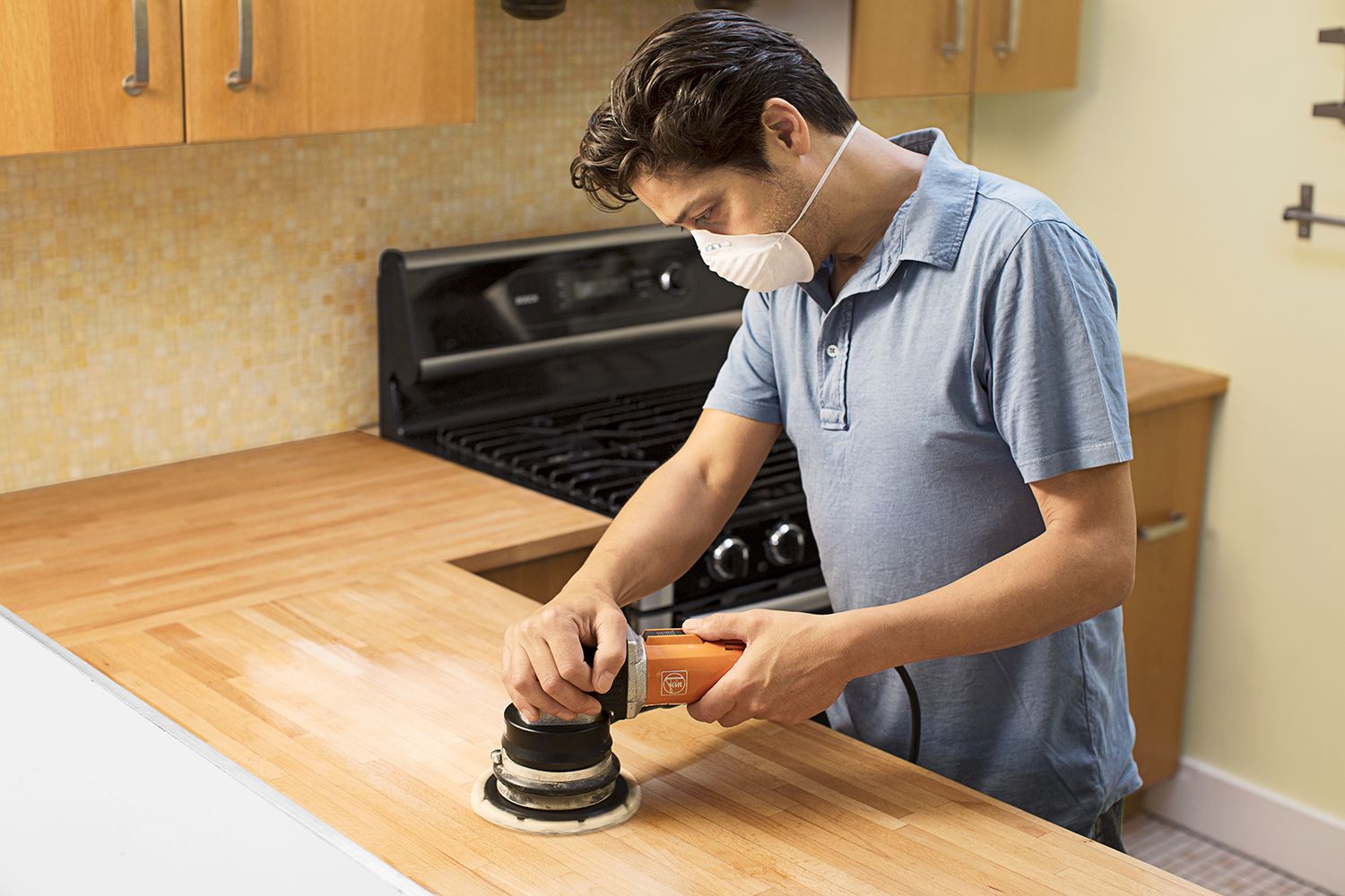 A man wearing a mask and using an electric sander on the surface of a butcher block countertop.