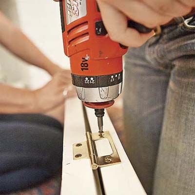 A person uses a drill to attach hinges to shutters so they can be used as cabinet doors.