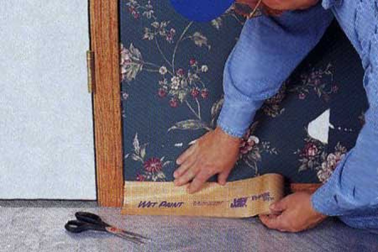 A man preparing to strip wallpaper by applying protective paper to a walls baseboard.