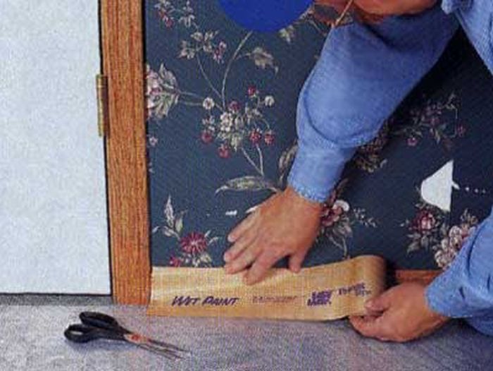 A man preparing to strip wallpaper by applying protective paper to a walls baseboard.