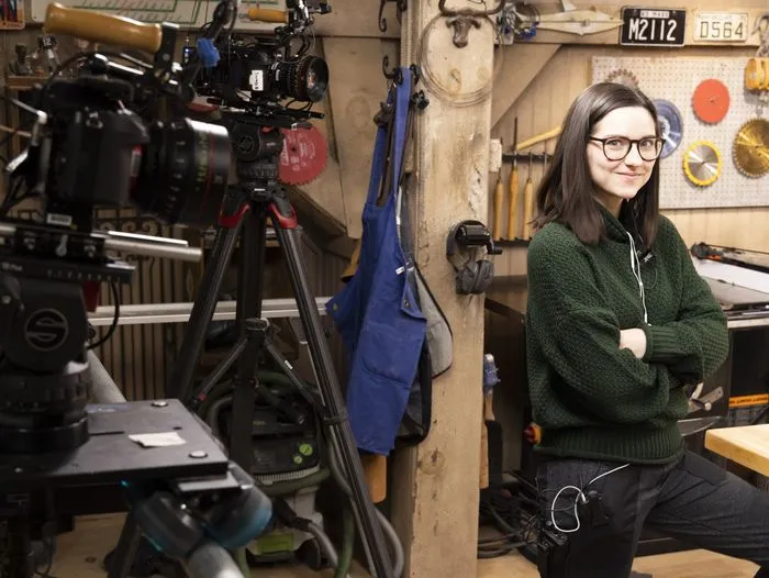 Sarah Chasse, wearing glasses and a green sweater, stands confidently with arms crossed in a workshop filled with tools, equipment, and cameras. She is ready for filming, with a camera tripod set up in the foreground.