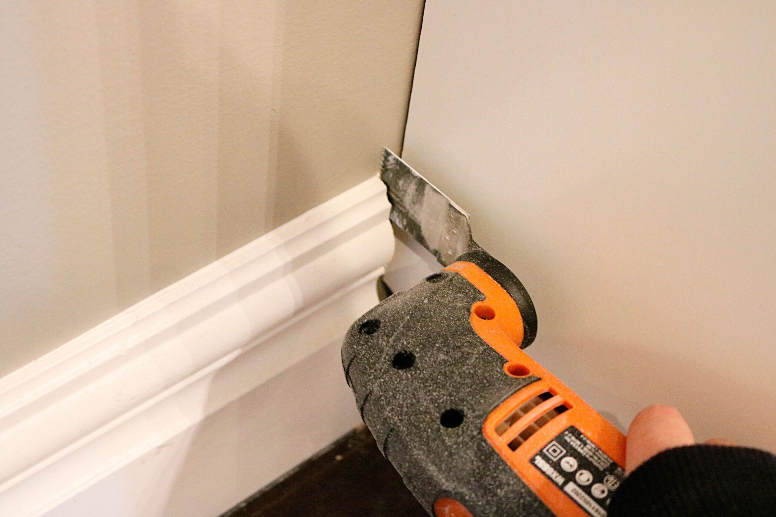 An oscillating tool with a flat blade being used to trim and remove a section of white baseboard trim along a wall where the built-in Ikea bookcase will go.