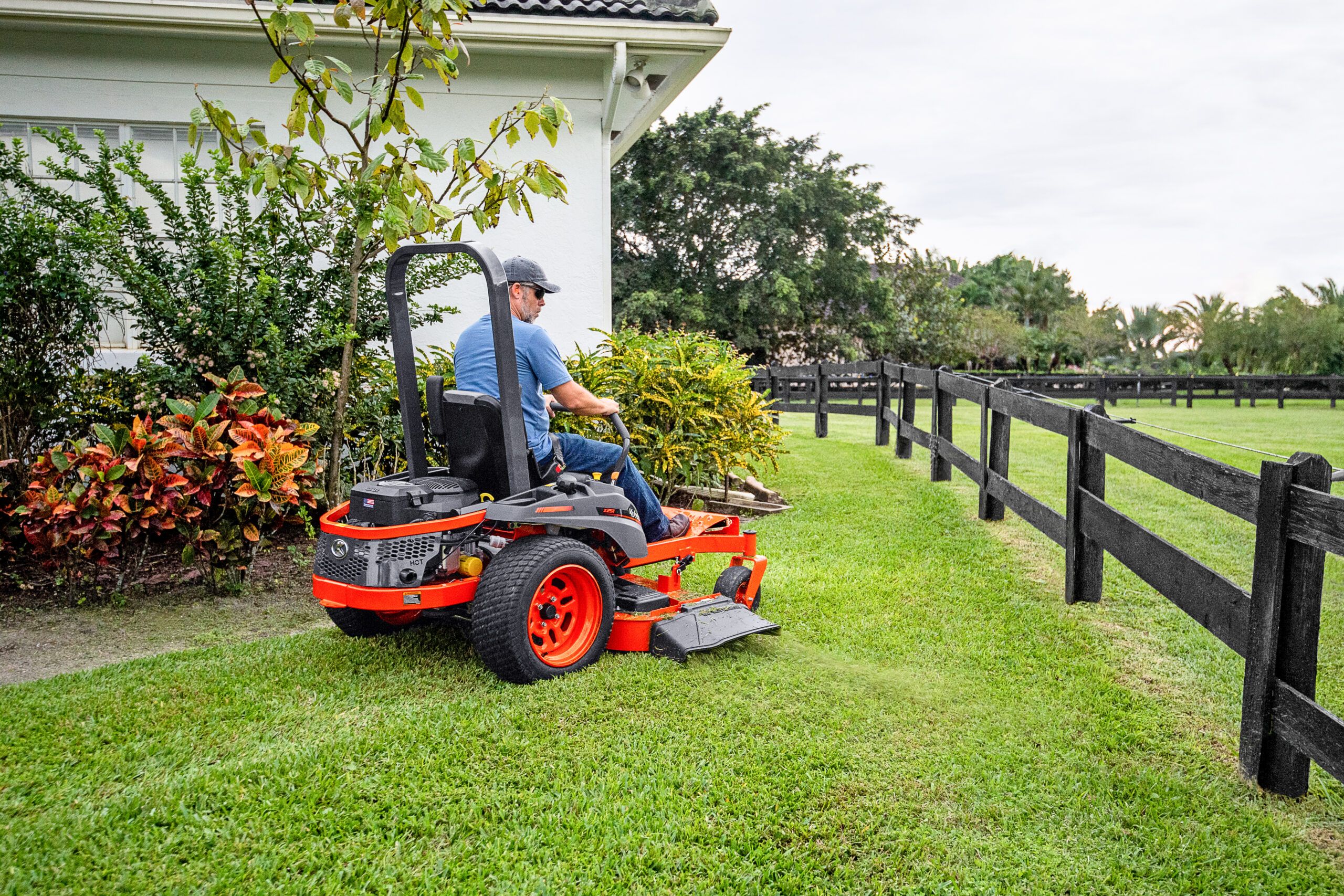 A man driving a zero-turn lawnmower beside some plants and a fence.