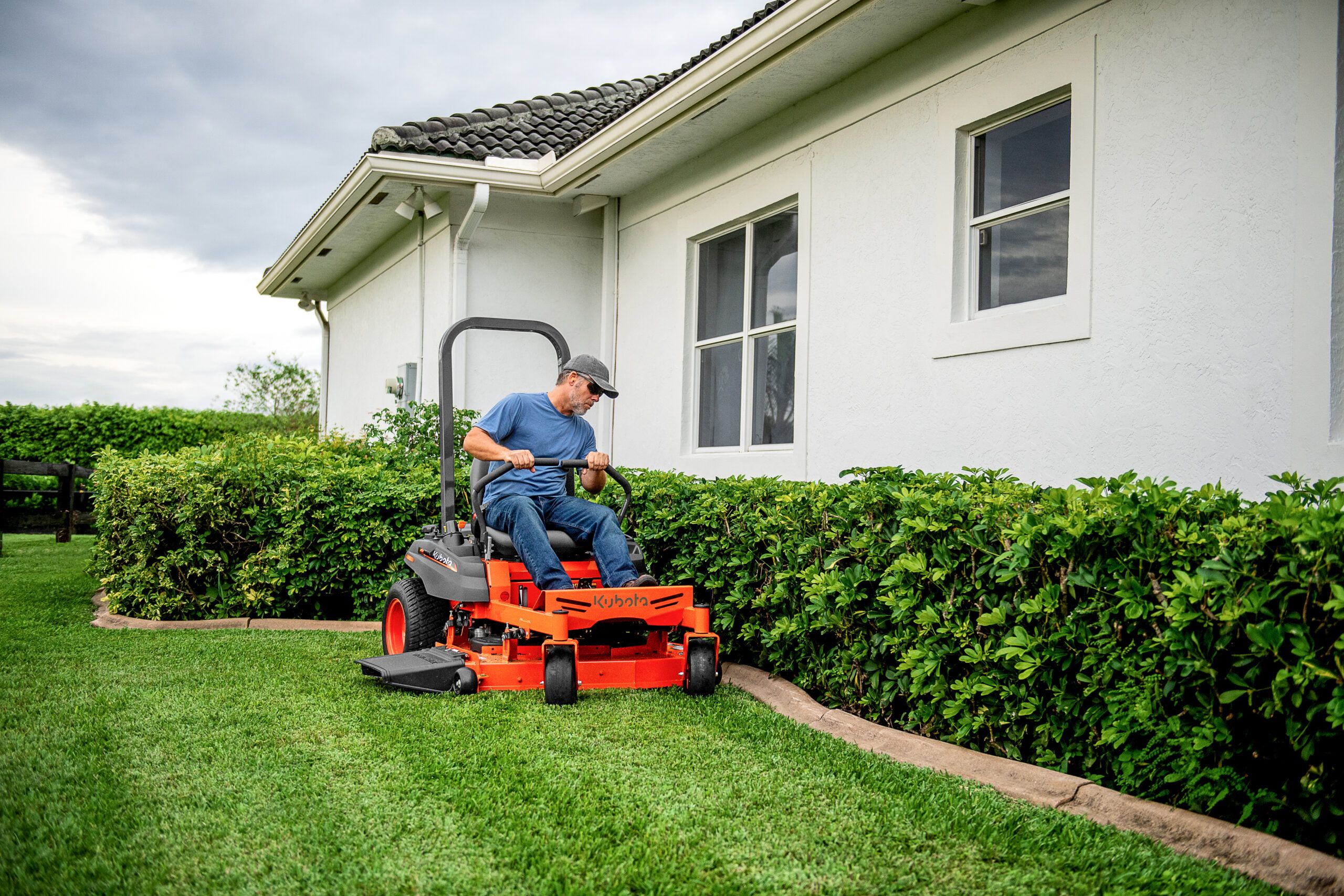 A man driving a zero-turn lawnmower next to a garden.