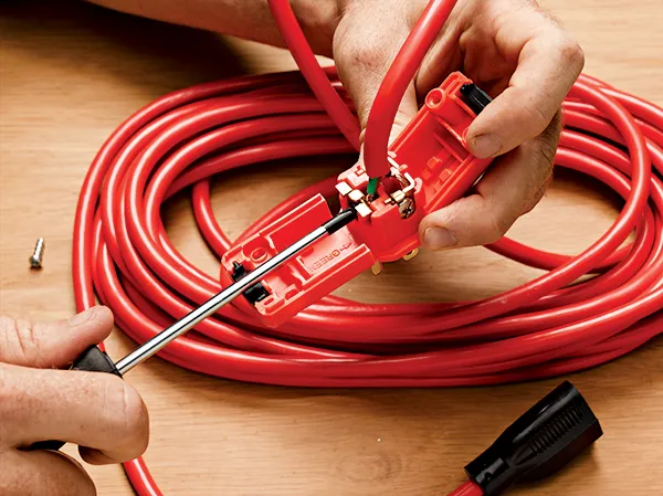 Hands using a screwdriver to connect wires inside the plug of a red extension cord, with the coiled cord in the background on a wooden surface.