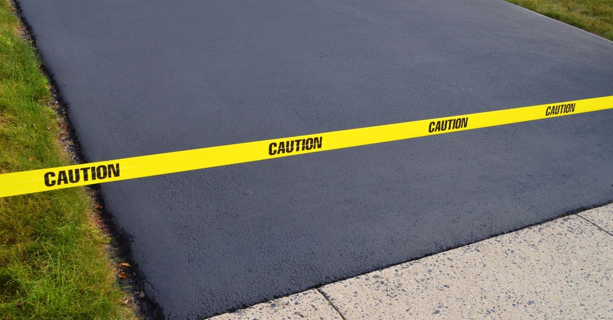 A freshly paved driveway with pristine sealcoat is marked with yellow caution tape. Lush green grass borders the sleek black asphalt on one side, while a light-colored concrete sidewalk runs along the bottom.