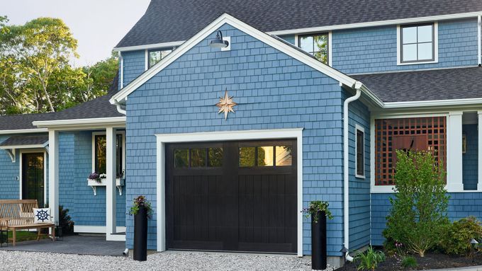 A blue house with white trim and a dark garage door boasts a decorative star above it. The gravel driveway is bordered by greenery, while tall black planters stand beside the garage, hinting at its all-about-cedar-shingles charm.