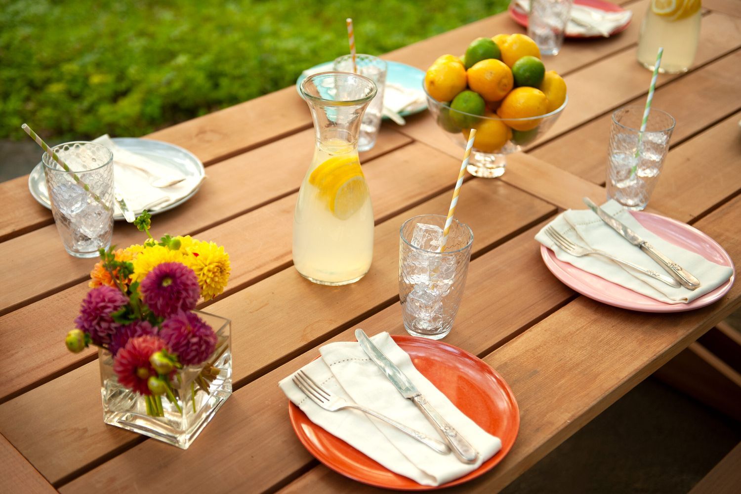 A wooden picnic table, crafted as one of the 4 DIY outdoor projects you can make out of real cedar, is set with colorful plates, napkins, and cutlery. Glasses with straws, a pitcher of lemonade, a bowl of lemons and limes, and vibrant flowers complete the cheerful setting.