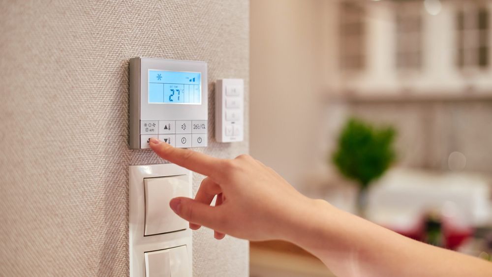 Lead image for the best smart thermostats guide, shows a hand reaching out to adjust the settings on white smart thermostat installed on a tan wall.