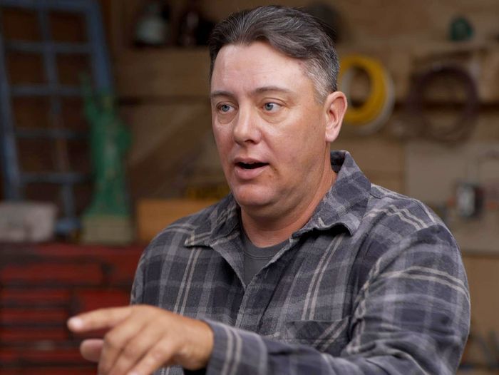 A man in a flannel shirt pointing towards the camera.