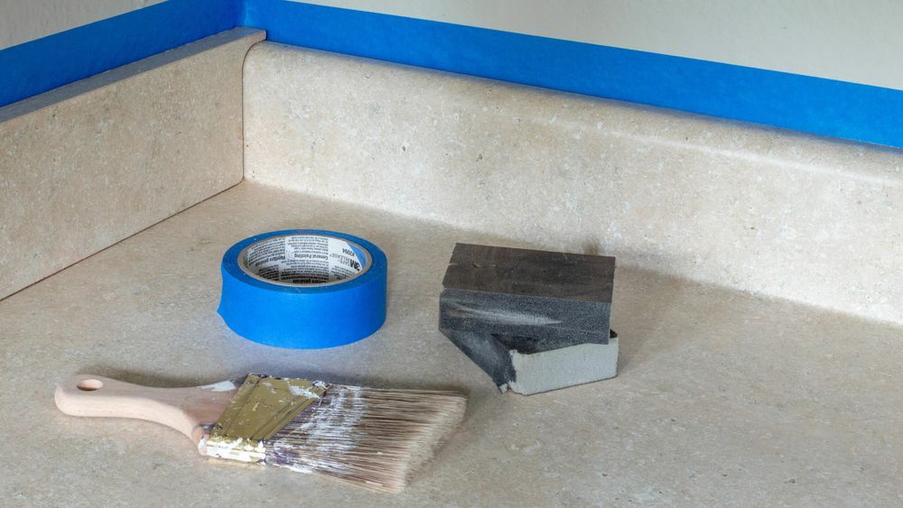 The supplies needed to prepare a laminate countertop, including a paint brush and blue tape.